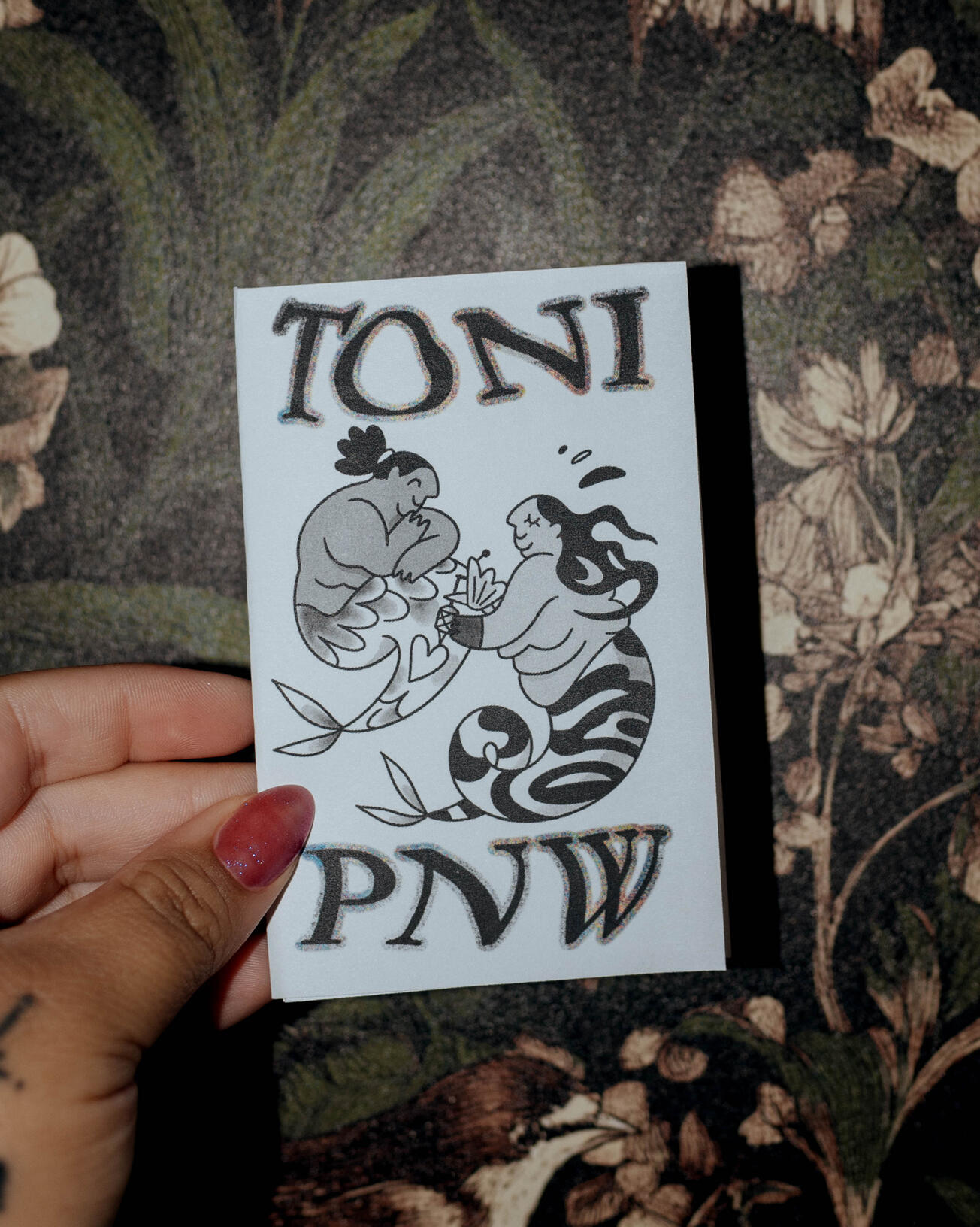 A photograph of a paper mini zine being held by a hand with purple nail polish against a dark, floral background. The zine cover says "TONI PNW" and features a digital drawing of two mermaids getting and giving a tattoo.