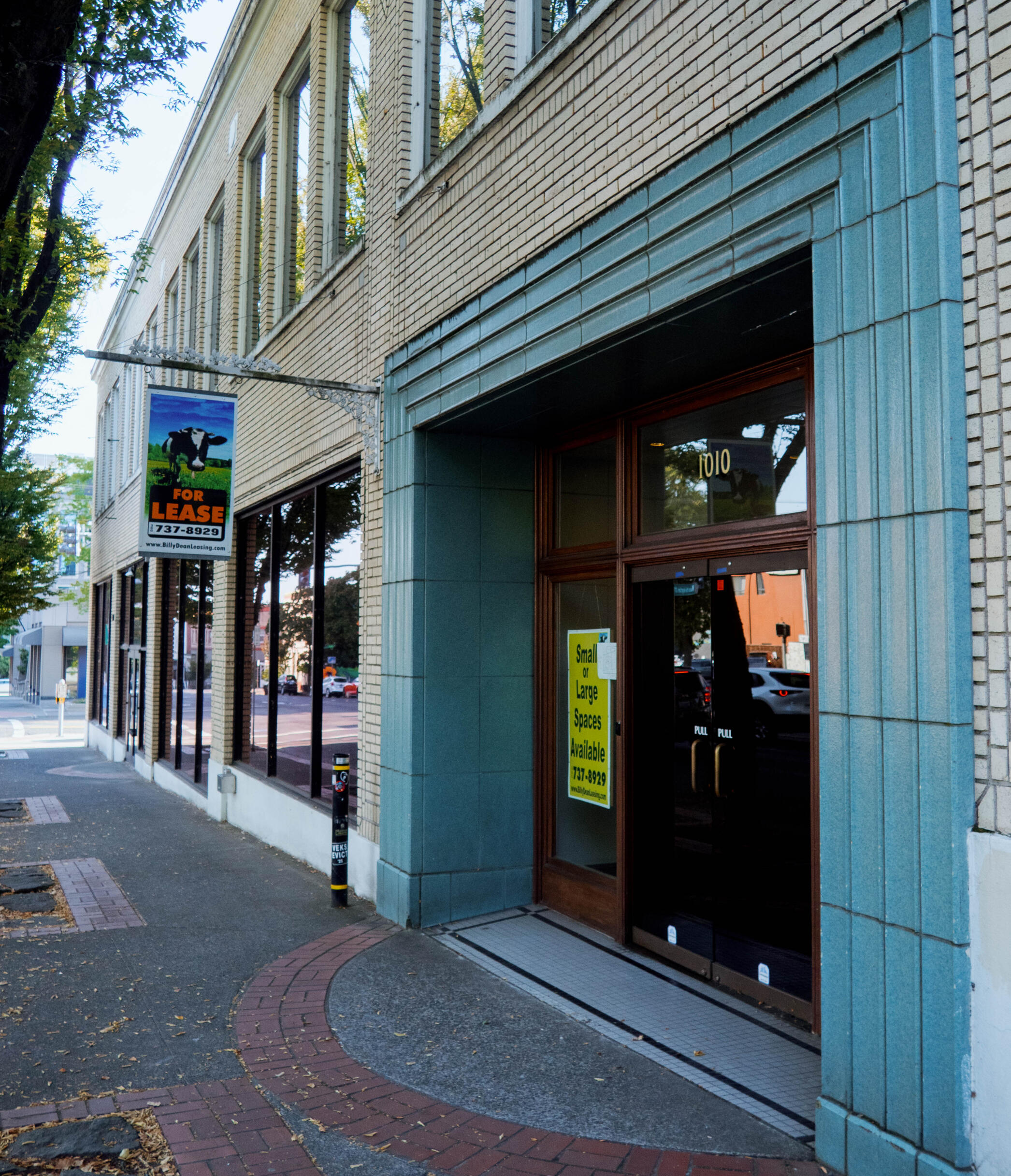 A photograph of the front doors of a two story beige building with a teal green archway above the main entrance, taken from the front sidewalk. Above the door are gold numbers "1010". In the window is a yellow sign saying "Small or Large Spaces Available 7